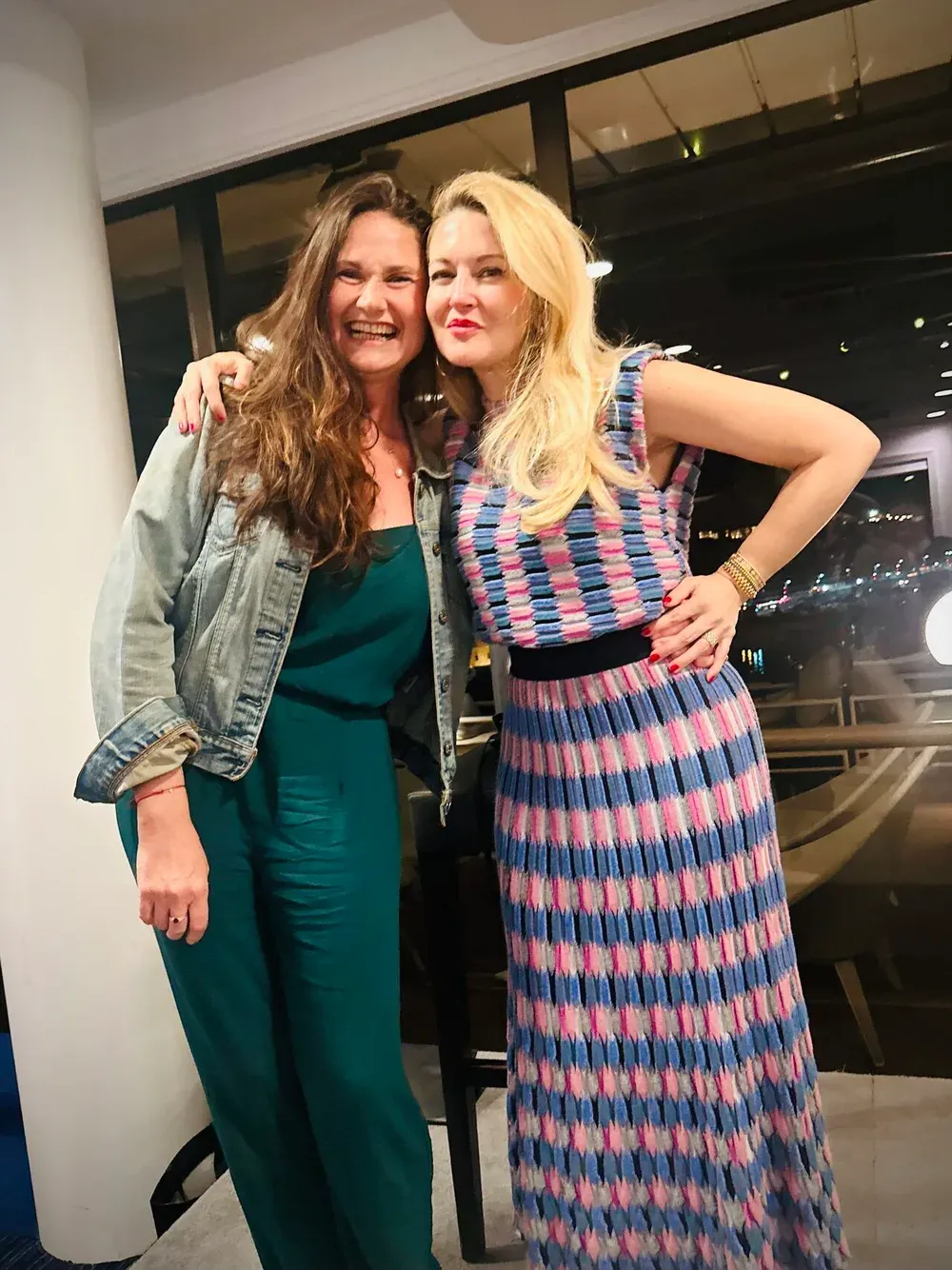 Deux femmes complices lors d’une soirée, la ville éclairée derrière les baies vitrées.