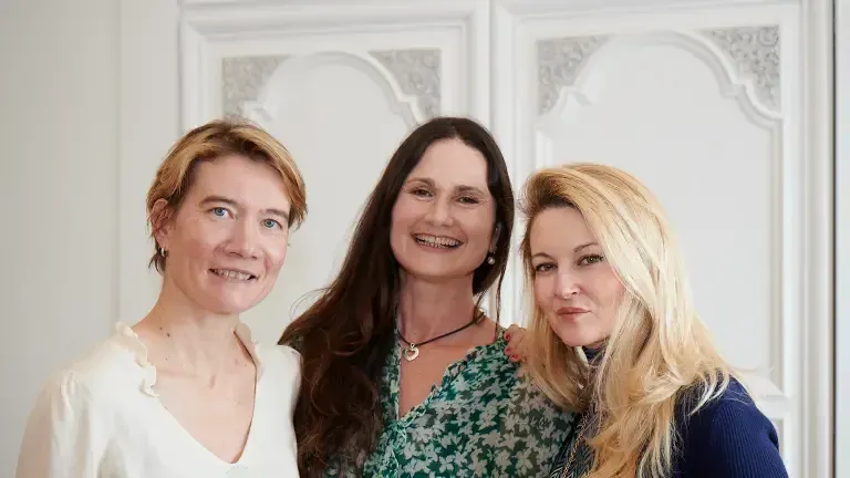 Trois femmes posent côte à côte devant une porte à moulures blanches.