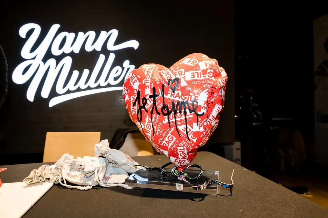 Un cœur sculpté recouvert d’adhésif « Fragile » et signé « je t’aime », posé devant le logo de l’artiste Yann Muller.