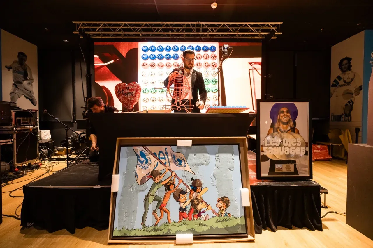 Sur la scène, l’animateur tourne le boulier devant l’écran des numéros ; à côté, les lots à gagner, dont un tableau et un disque d’or.