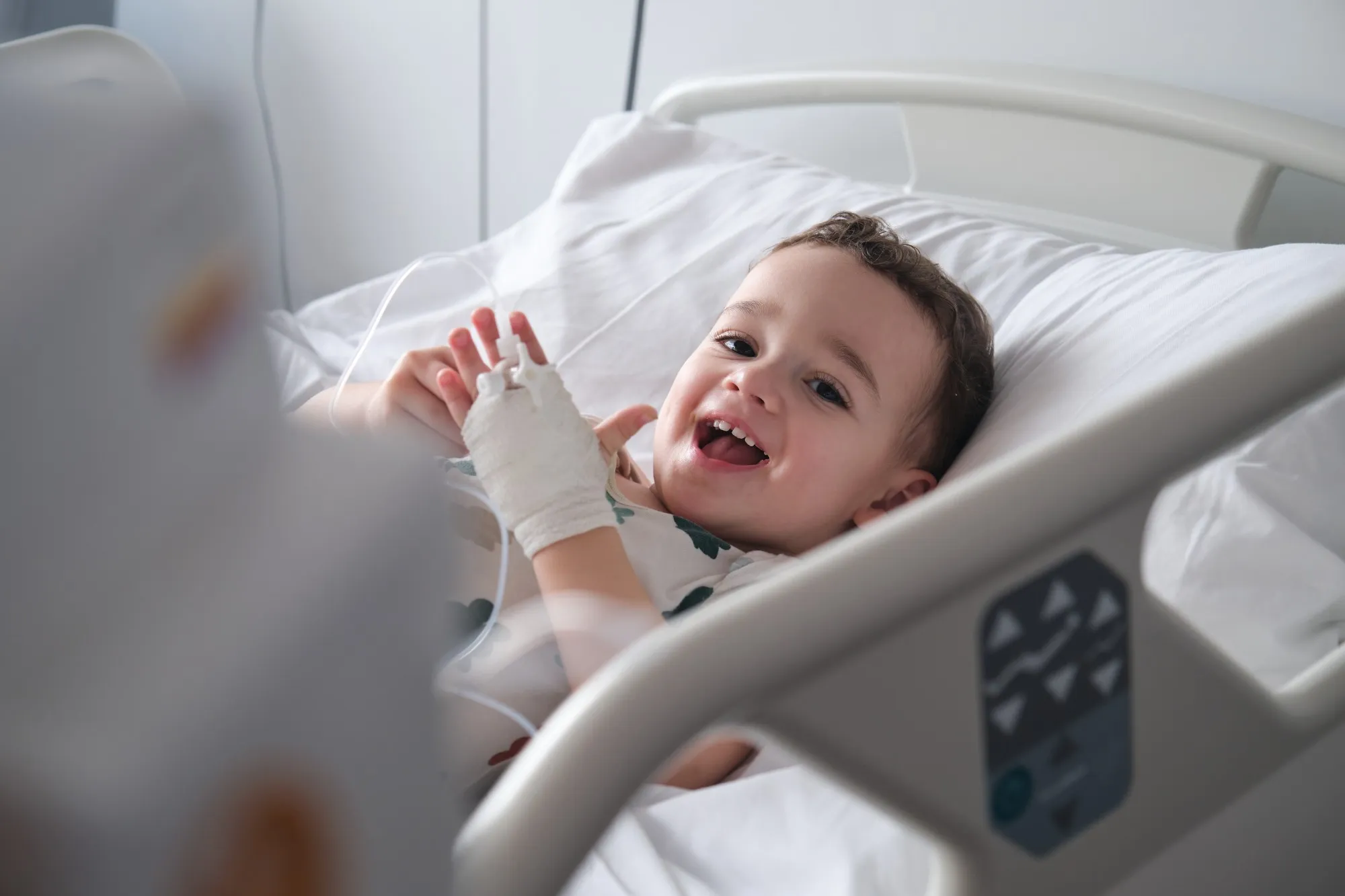 Un jeune enfant hospitalisé, sourire aux lèvres, la main pansée dans son lit d’hôpital.
